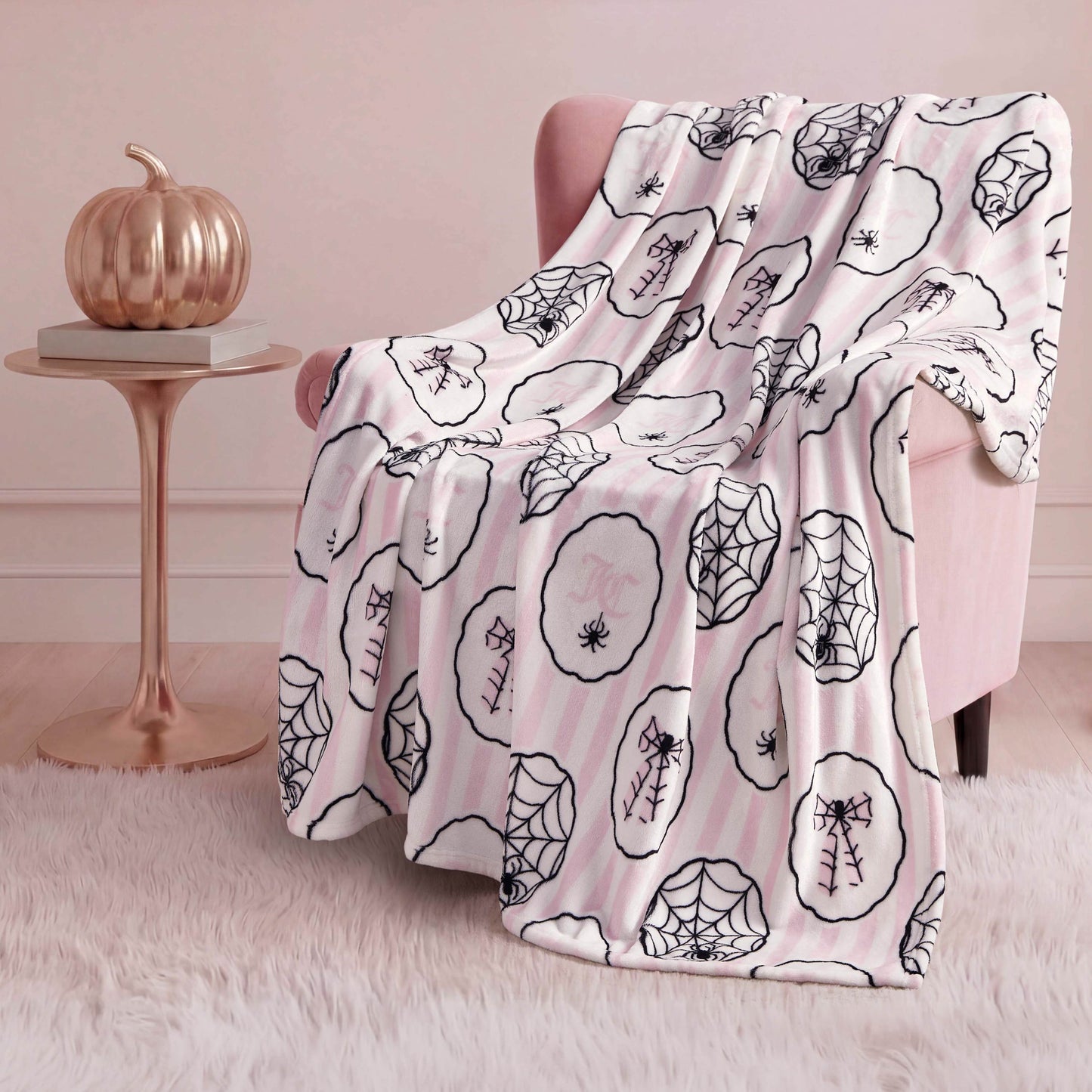 A pink armchair is draped with a Halloween Plush Throw Blanket 50" x 60" featuring black spiderweb and spider designs. Beside it, a small round table holds a metallic pumpkin, all set on a white fluffy rug.