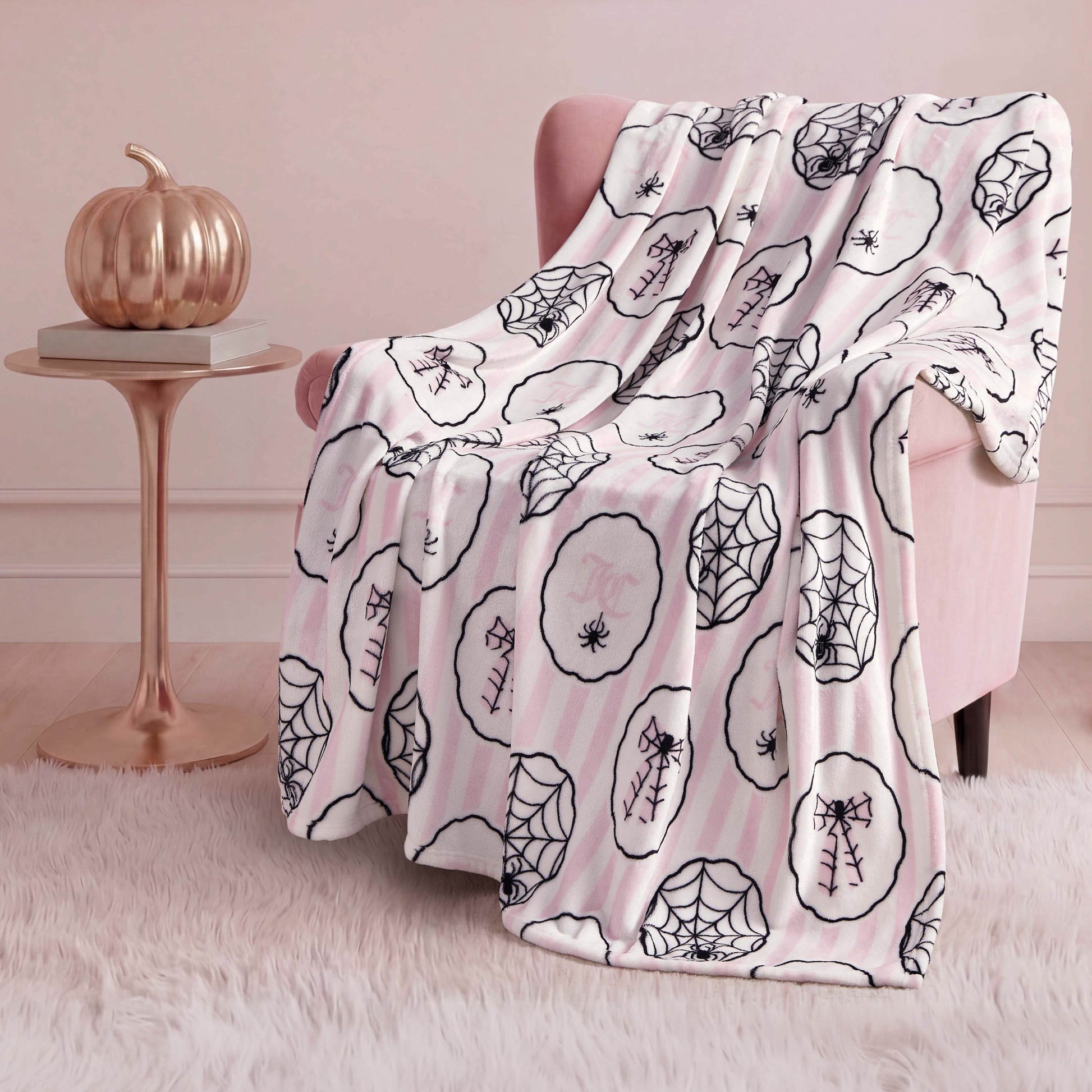 A pink armchair is draped with a Halloween Plush Throw Blanket 50" x 60" featuring black spiderweb and spider designs. Beside it, a small round table holds a metallic pumpkin, all set on a white fluffy rug.