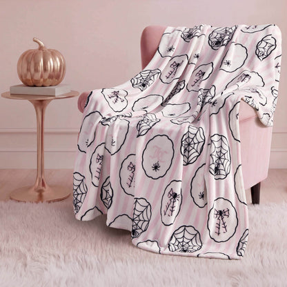 A pink armchair is draped with a Halloween Plush Throw Blanket 50" x 60" featuring black spiderweb and spider designs. Beside it, a small round table holds a metallic pumpkin, all set on a white fluffy rug.