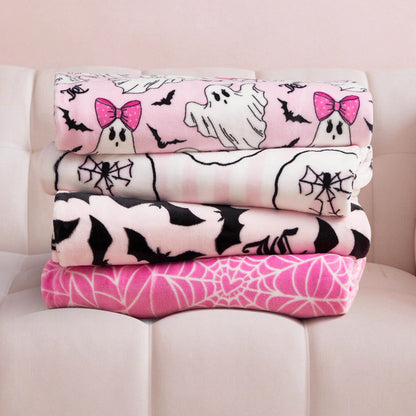 Four folded Halloween Plush Throw Blankets (50" x 60") with ghost, bat, and spider designs are stacked on a white sofa against a light pink wall—perfect for adding cozy Juicy Couture style to your space.