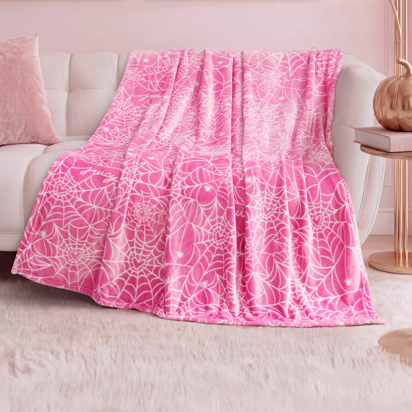 The Halloween Plush Throw Blanket 50" x 60" in pink with a white web pattern is draped over a white couch in a pastel-themed room with a plush white rug, pink pillow, and copper lamp—adding cozy Halloween décor.