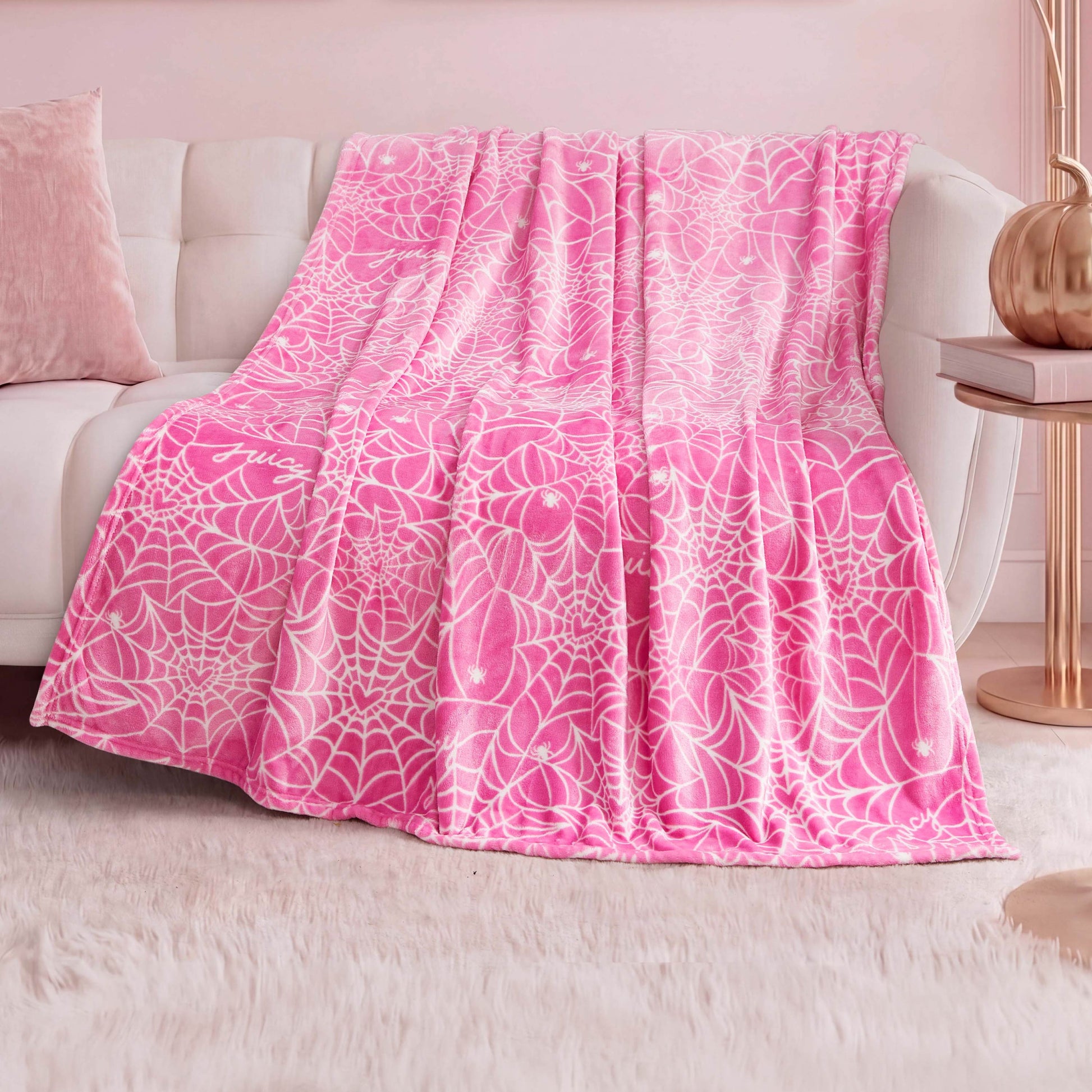 The Halloween Plush Throw Blanket 50" x 60" in pink with a white web pattern is draped over a white couch in a pastel-themed room with a plush white rug, pink pillow, and copper lamp—adding cozy Halloween décor.