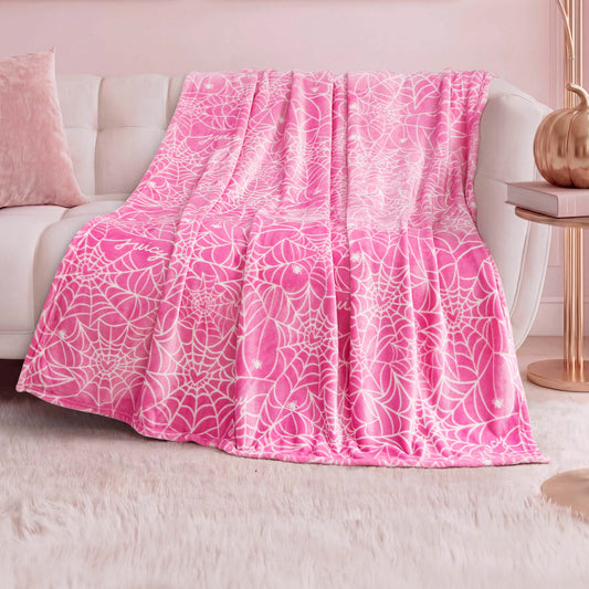 The Halloween Plush Throw Blanket 50" x 60" in pink with a white web pattern is draped over a white couch in a pastel-themed room with a plush white rug, pink pillow, and copper lamp—adding cozy Halloween décor.