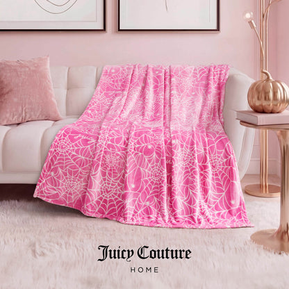 A Juicy Couture Halloween Plush Throw Blanket 50" x 60" in pink and white is draped over a white sofa in a modern living room with pink décor—lamp, art, pillow, and rug. "Juicy Couture Home" is displayed at the bottom.