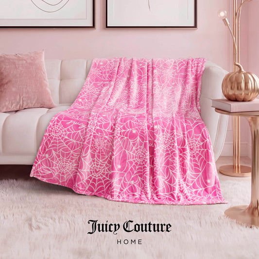 A Juicy Couture Halloween Plush Throw Blanket 50" x 60" in pink and white is draped over a white sofa in a modern living room with pink décor—lamp, art, pillow, and rug. "Juicy Couture Home" is displayed at the bottom.