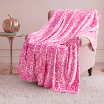 The Halloween Plush Throw Blanket 50" x 60" in soft pink with a white web pattern is draped over a pink armchair beside a round table with a metallic pumpkin, all atop a white furry rug in a cozy room.