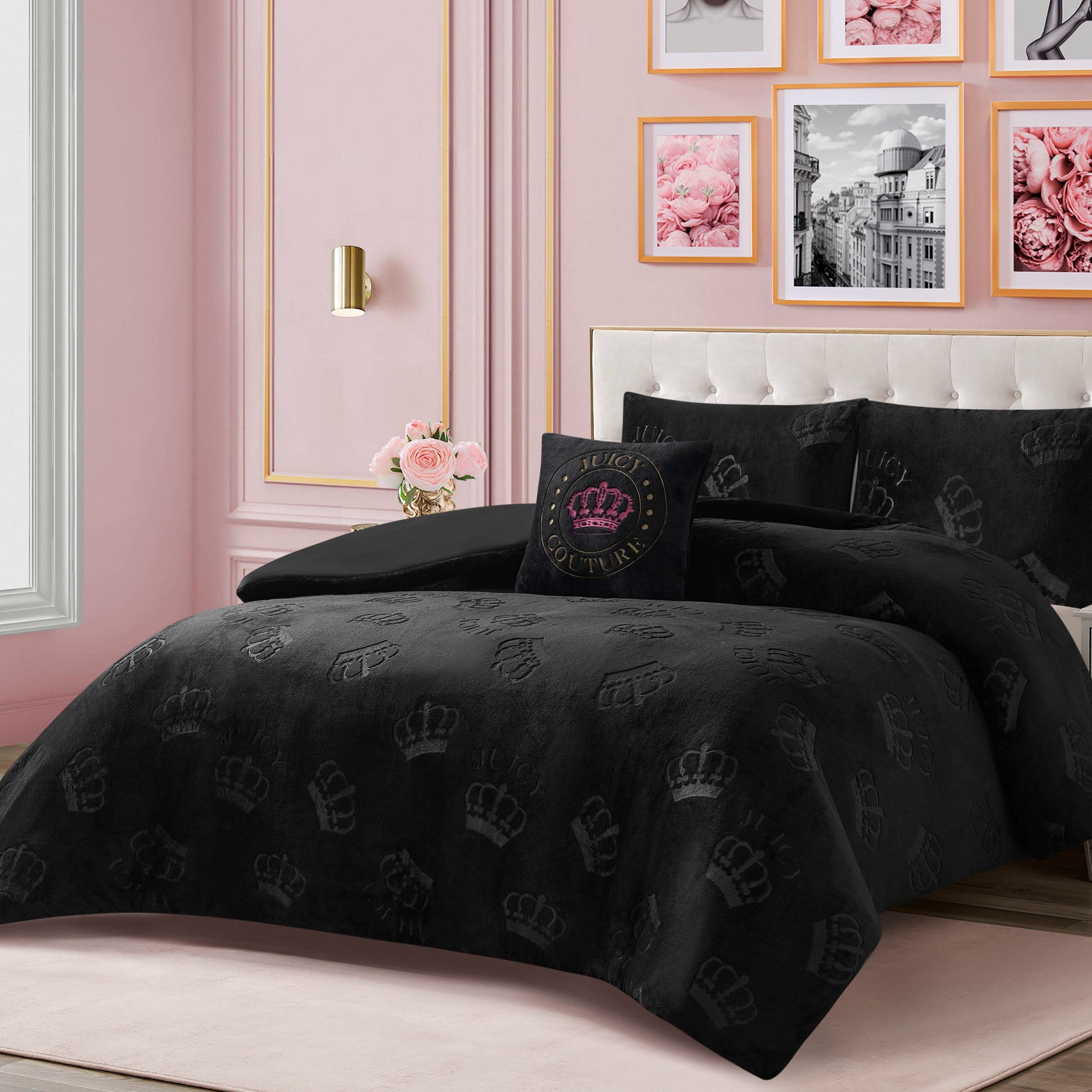 A neatly made bed with glamorous bedding, featuring the Embossed Plush Crown Comforter Set from Juicy Couture and a Juicy Couture crown pillow, stands out against pink walls adorned with framed floral and city photos. A vase of pink roses on the nightstand adds an extra touch of elegance.