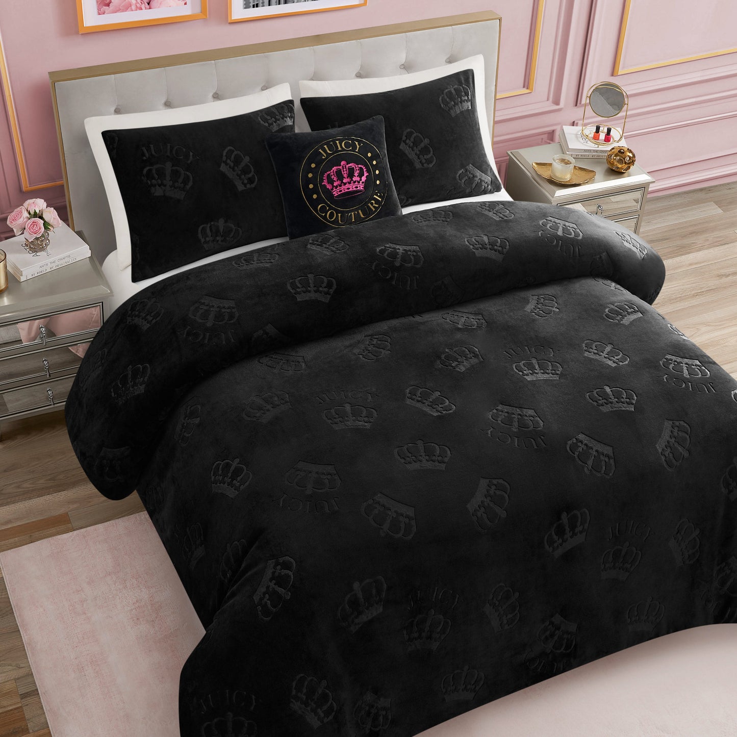 A neatly made bed showcases the Embossed Plush Crown Comforter Set by Juicy Couture in black, featuring subtle crown patterns, matching black pillows, and a decorative black pillow with a crest logo. The pink walls and mirrored nightstands complete the glamorous bedding look.