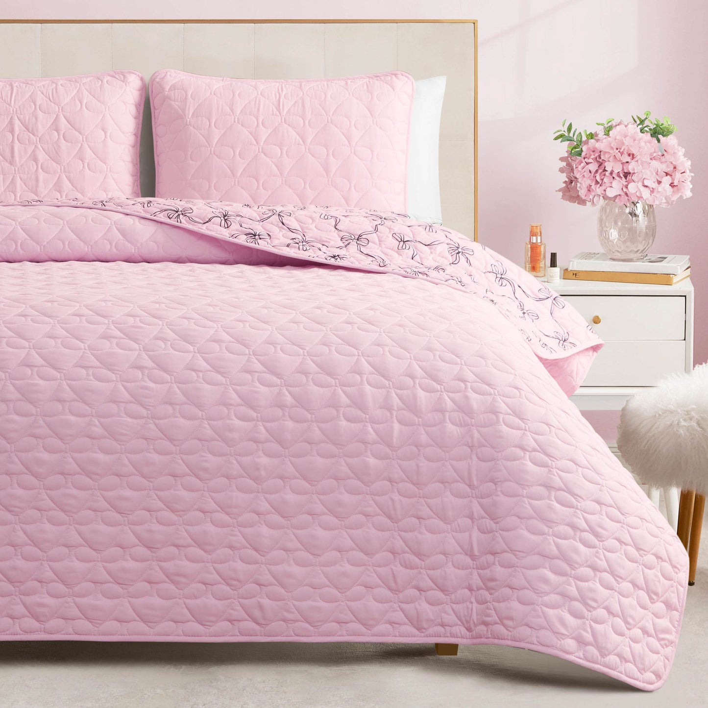 A bed featuring the Luxe Bow Reversible Quilt Set in light pink, complete with matching pillows, sits beside a white nightstand adorned with a vase of flowers and a small bottle, while a fluffy white stool adds charm to the softly lit room.