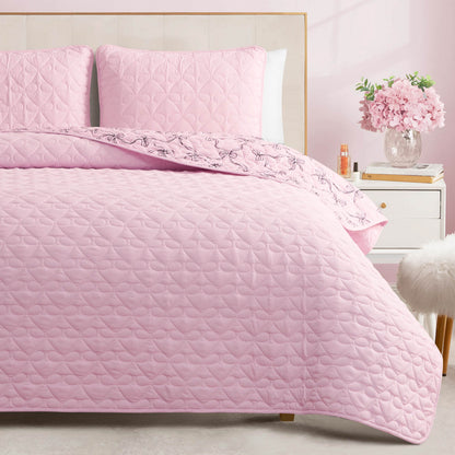 A bed featuring the Luxe Bow Reversible Quilt Set in light pink, complete with matching pillows, sits beside a white nightstand adorned with a vase of flowers and a small bottle, while a fluffy white stool adds charm to the softly lit room.