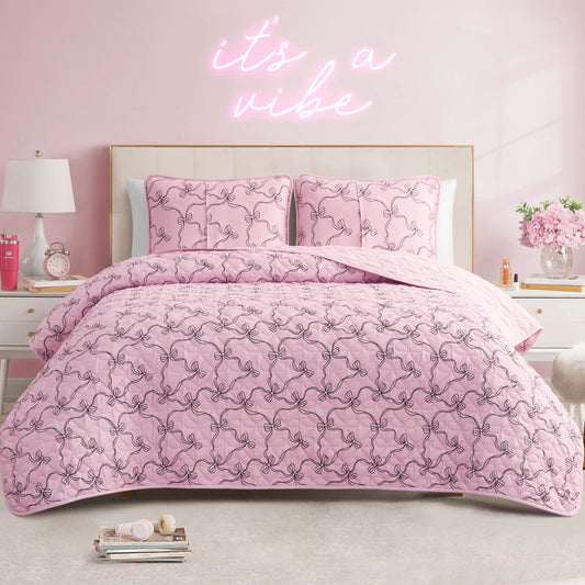 A pink bedroom with a bed covered in the Luxe Bow Reversible Quilt Set, featuring a pink patterned comforter and matching pillows. A neon sign on the wall reads "it's a vibe," while nightstands with decor and flowers sit on each side of the bed.