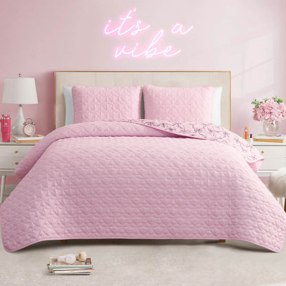 A neatly made bed with a Luxe Bow Reversible Quilt Set and pink pillows stands in a pastel pink bedroom. A neon sign above the bed reads "it's a vibe." Decorative items, flowers, and lamps sit on the white nightstands.