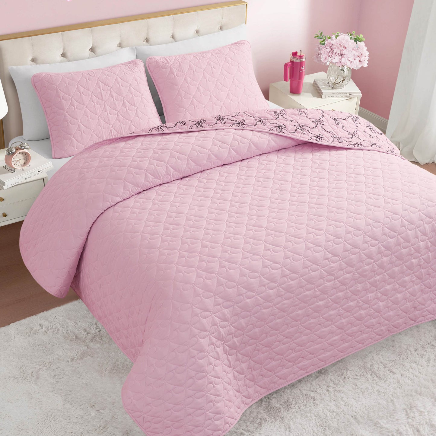 A neatly made bed features the Luxe Bow Reversible Quilt Set and matching pillows in a pink-themed bedroom. Flowers, a pink lamp, and a nightstand sit next to the bed, while a rug covers part of the wooden floor.