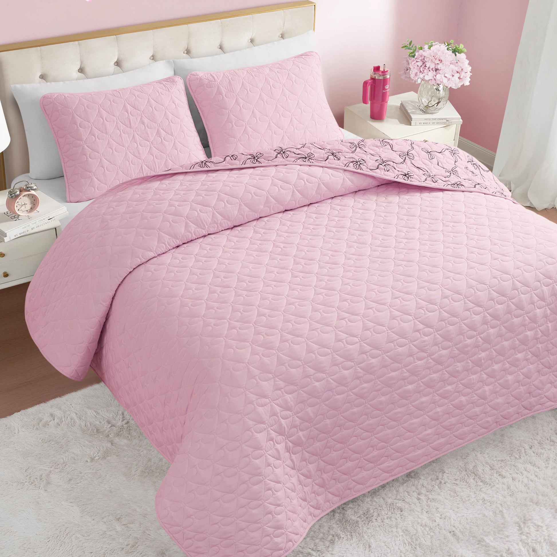 A neatly made bed features the Luxe Bow Reversible Quilt Set and matching pillows in a pink-themed bedroom. Flowers, a pink lamp, and a nightstand sit next to the bed, while a rug covers part of the wooden floor.