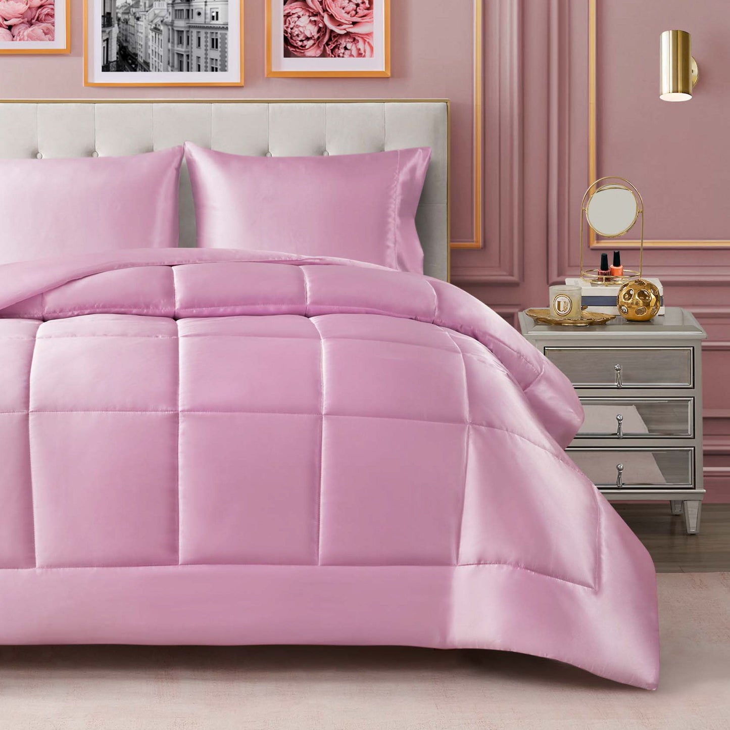 A neatly made bed features the Solid Satin Down Alternative Comforter in a shiny pink satin finish, paired with matching pillows beside a nightstand holding a mirror, clock, and decor. Pink wall panels, framed photos, and a gold sconce complete the chic bedroom decor while providing all-season warmth.