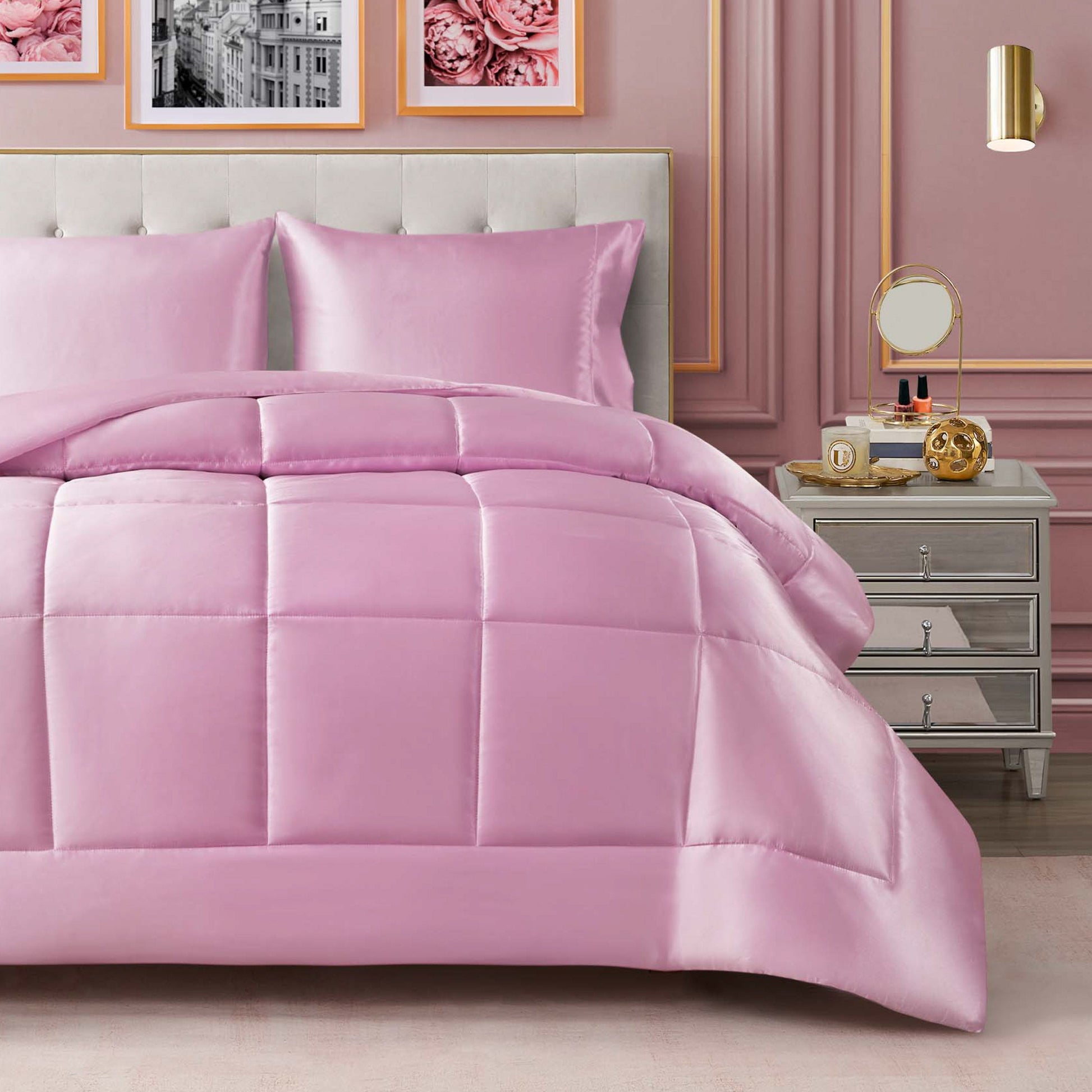 A neatly made bed features the Solid Satin Down Alternative Comforter in a shiny pink satin finish, paired with matching pillows beside a nightstand holding a mirror, clock, and decor. Pink wall panels, framed photos, and a gold sconce complete the chic bedroom decor while providing all-season warmth.