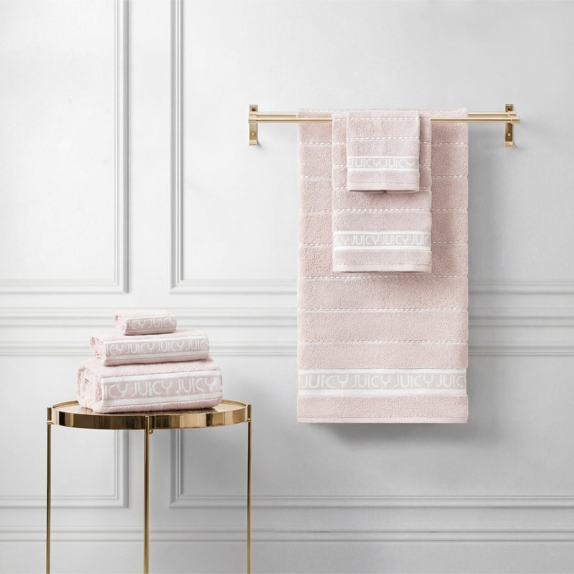 A set of Plush Stripe Bath Towel Set towels in pale pink is neatly arranged—some folded on a round gold table, others hanging on a gold towel bar against a white paneled wall.