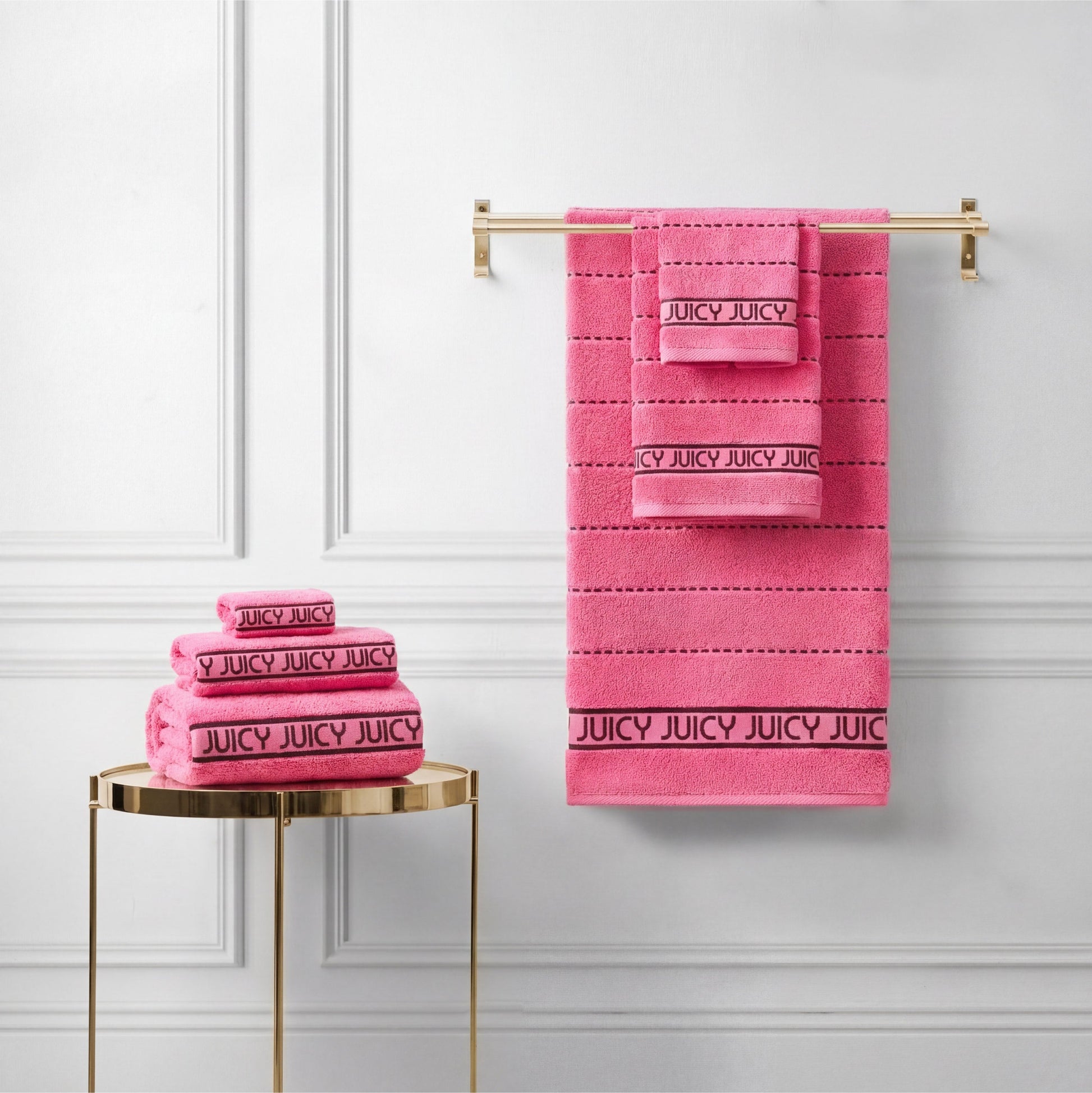 A Plush Stripe Bath Towel Set in bright pink, featuring "JUICY" on black bands, is arranged on a gold rack and round gold side table against a white paneled wall.