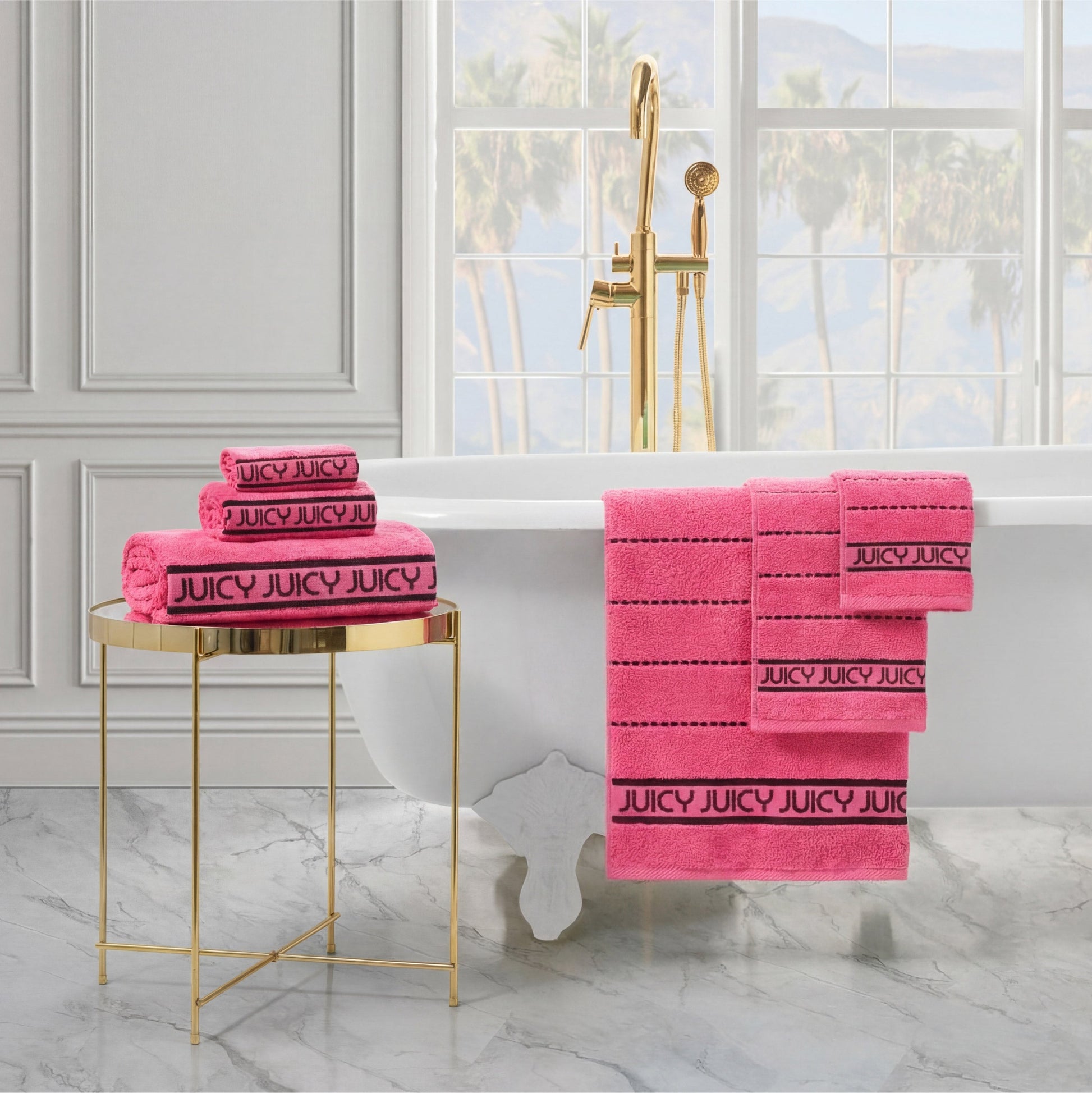 Plush Stripe Bath Towel Set in bright pink is neatly folded and hanging in a luxurious bathroom featuring a white clawfoot tub, gold fixtures, a round gold side table, and a window with palm trees outside.