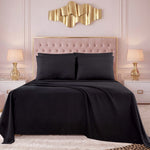 A bed dressed in the Microfiber Sheet Set With Extra Pillowcases in black, accented with two pillows, is positioned between two nightstands topped with lamps. The headboard is tufted, and a gold abstract wall art piece hangs above it. Soft pink walls and a light-colored rug complete the room’s look.