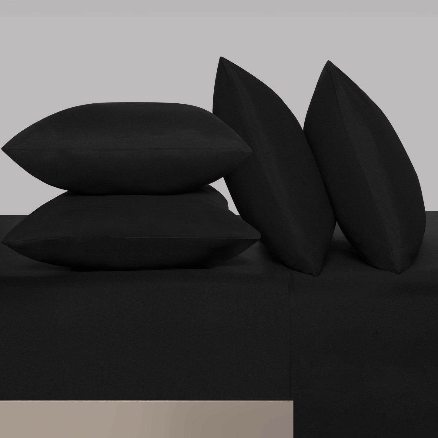 Four black pillows are arranged on a matching black Microfiber Sheet Set With Extra Pillowcases, with two pillows stacked horizontally and two positioned upright in the background, creating an elegant display of luxury bedding.