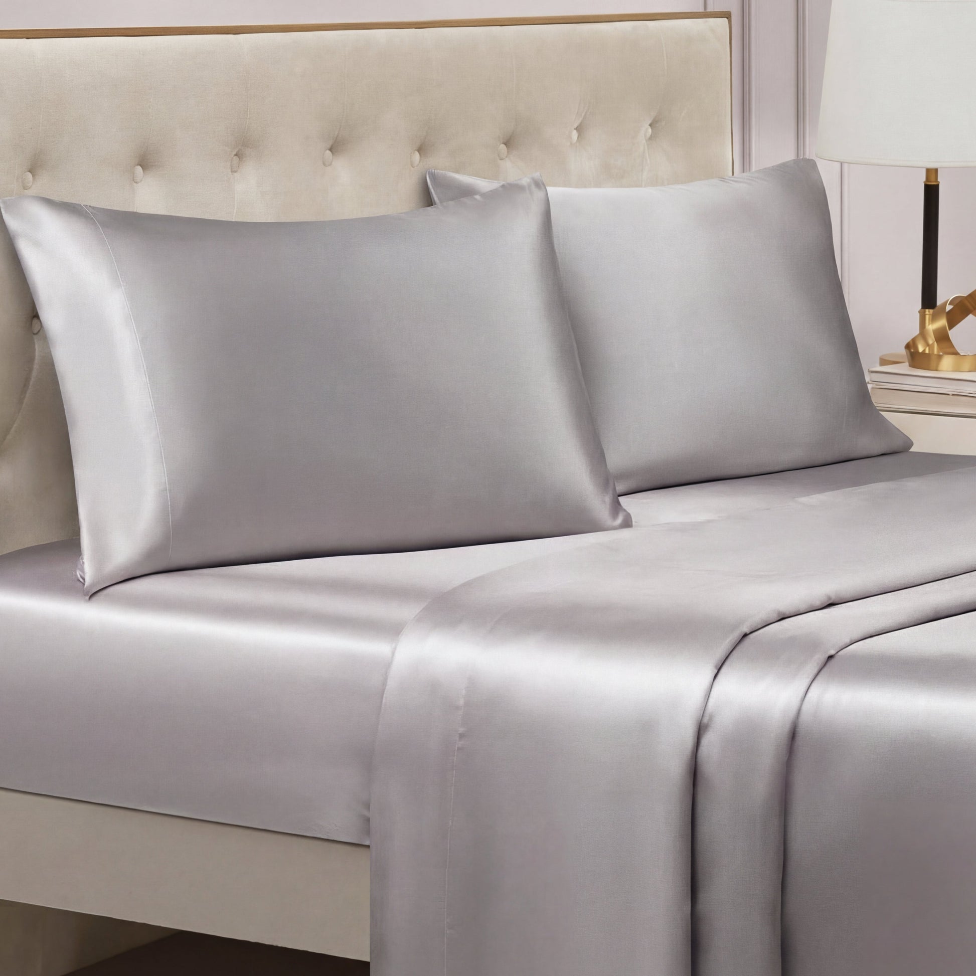 A bed with a tufted beige headboard features the Solid Satin Sheet Set in shiny silver-gray. On the bedside table in the background, a lamp and decorative object can also be seen.