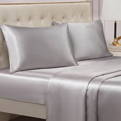 A bed with a tufted beige headboard features the Solid Satin Sheet Set in shiny silver-gray. On the bedside table in the background, a lamp and decorative object can also be seen.