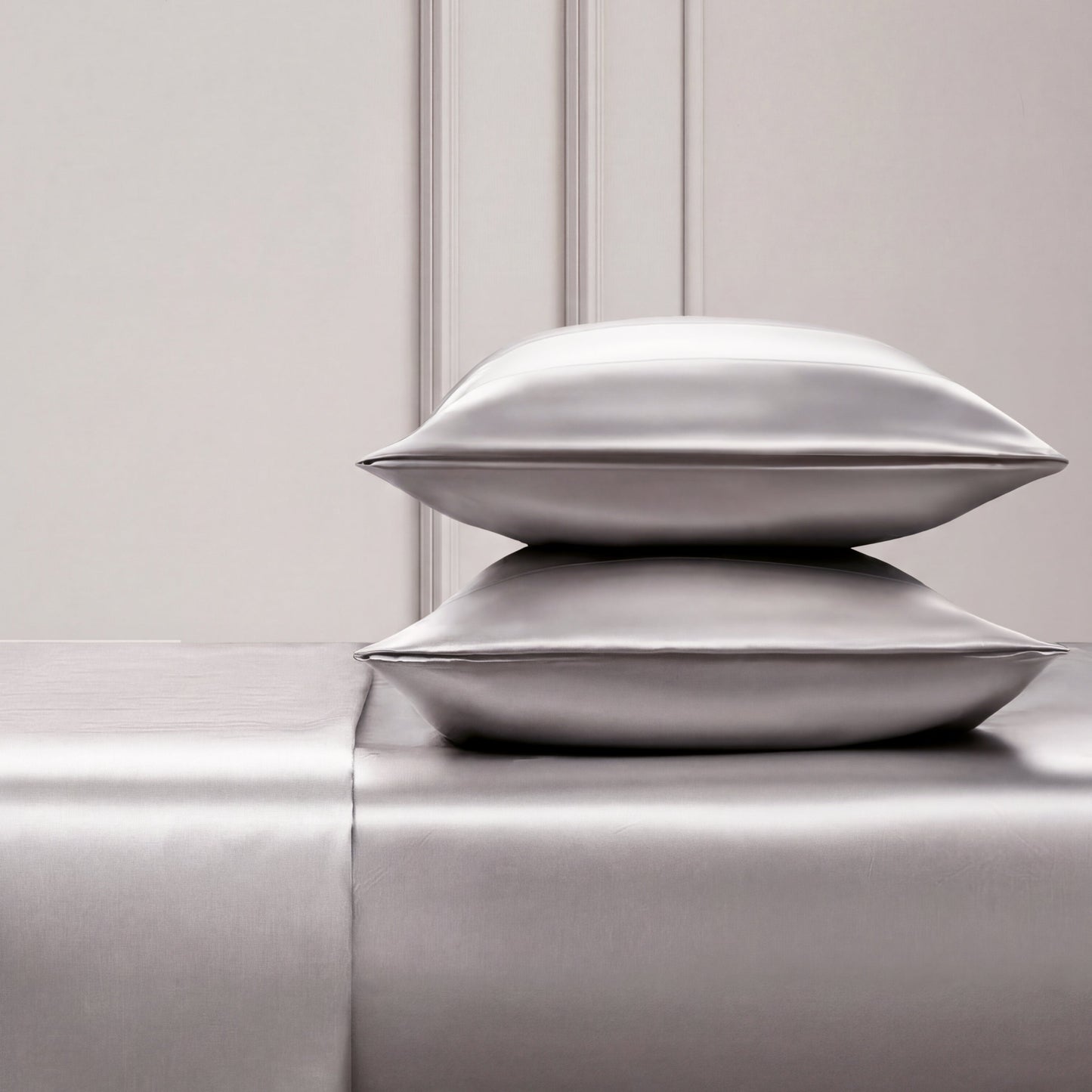 Two shiny silver satin pillows rest on a matching Solid Satin Sheet Set, placed against an elegant light-paneled wall. The scene exudes luxury and minimalism, ideal for those who love ultra-soft satin bedding.
