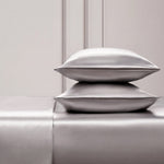 Two shiny silver satin pillows rest on a matching Solid Satin Sheet Set, placed against an elegant light-paneled wall. The scene exudes luxury and minimalism, ideal for those who love ultra-soft satin bedding.