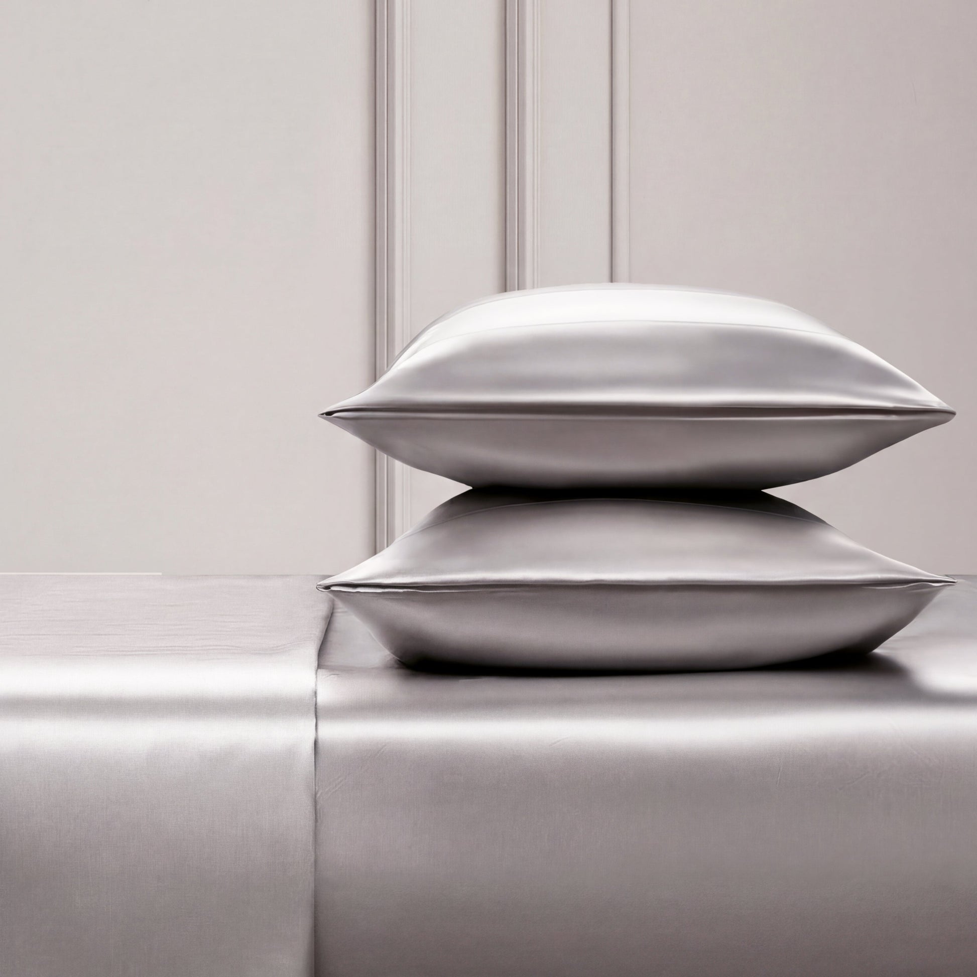 Two shiny silver satin pillows rest on a matching Solid Satin Sheet Set, placed against an elegant light-paneled wall. The scene exudes luxury and minimalism, ideal for those who love ultra-soft satin bedding.