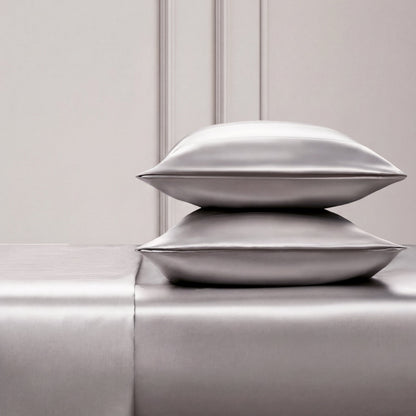 Two shiny silver satin pillows rest on a matching Solid Satin Sheet Set, placed against an elegant light-paneled wall. The scene exudes luxury and minimalism, ideal for those who love ultra-soft satin bedding.