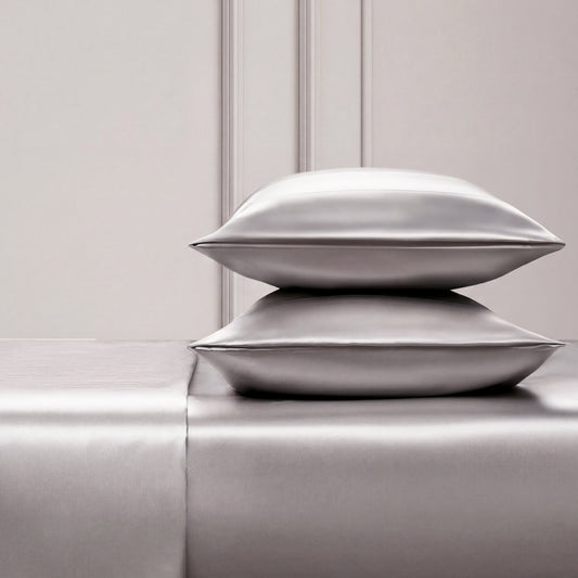 Two shiny silver satin pillows rest on a matching Solid Satin Sheet Set, placed against an elegant light-paneled wall. The scene exudes luxury and minimalism, ideal for those who love ultra-soft satin bedding.