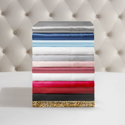 A stack of Solid Satin Sheet Sets in colors like grey, blue, white, pink, red, black, and leopard print rests on a white surface against a tufted light background.