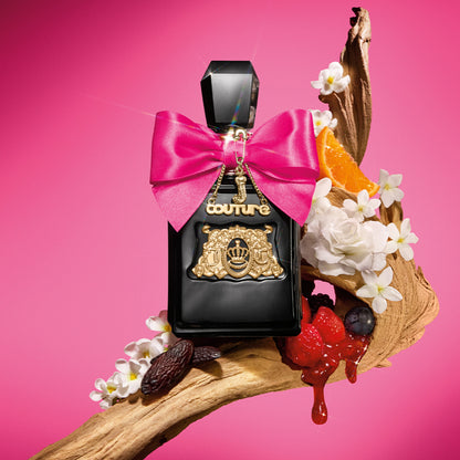 A Viva La Juicy Elixir Eau de Parfum Spray, 3.4 oz bottle in black with gold accents and a pink bow rests on a branch, surrounded by white flowers, an orange slice, berries, and syrup against a vibrant pink background.
