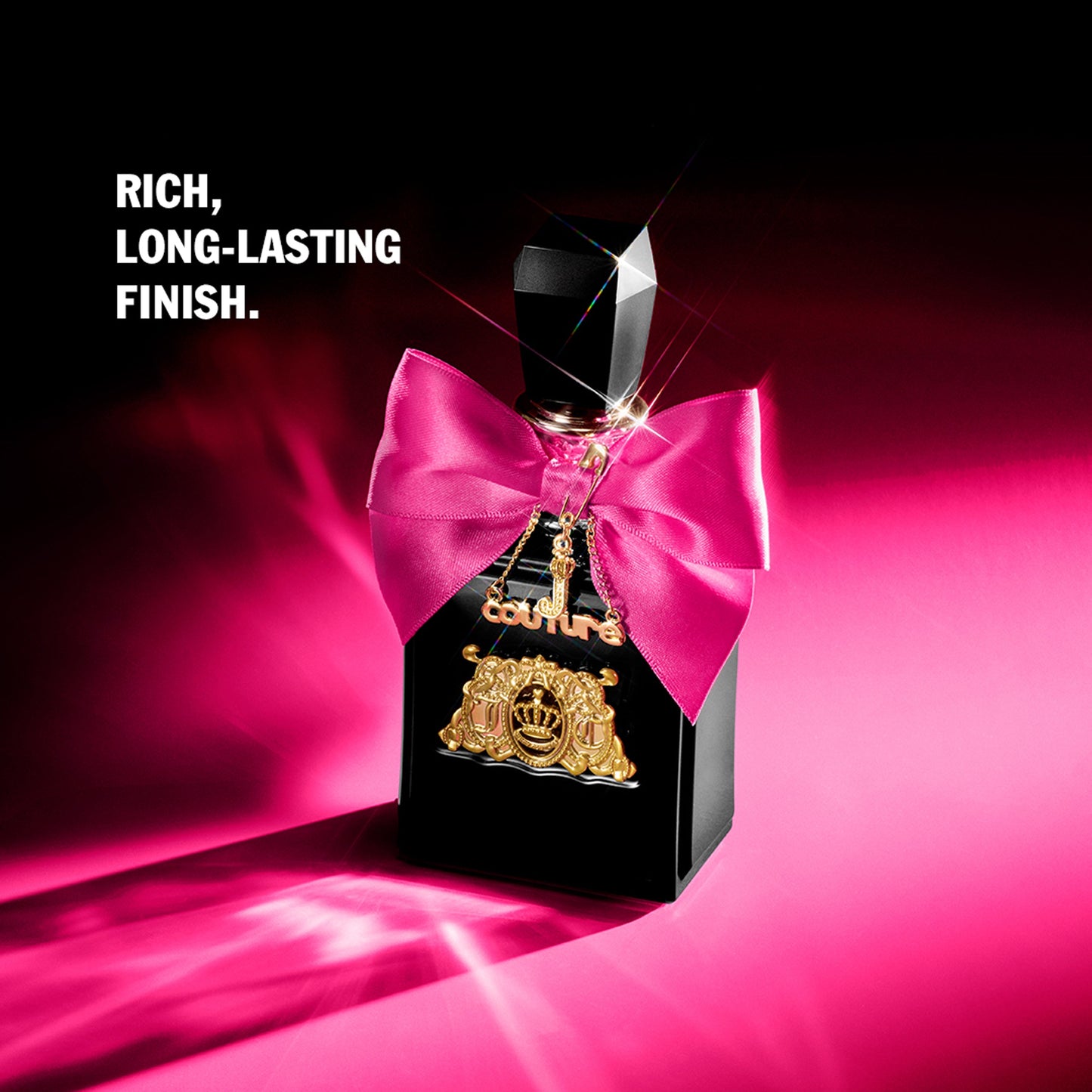 A bottle of Viva La Juicy Elixir Eau de Parfum Spray, 1.7 oz, adorned with a pink bow and gold charms, sits on a vivid pink surface under a spotlight. Text on the left highlights its rich, long-lasting finish.