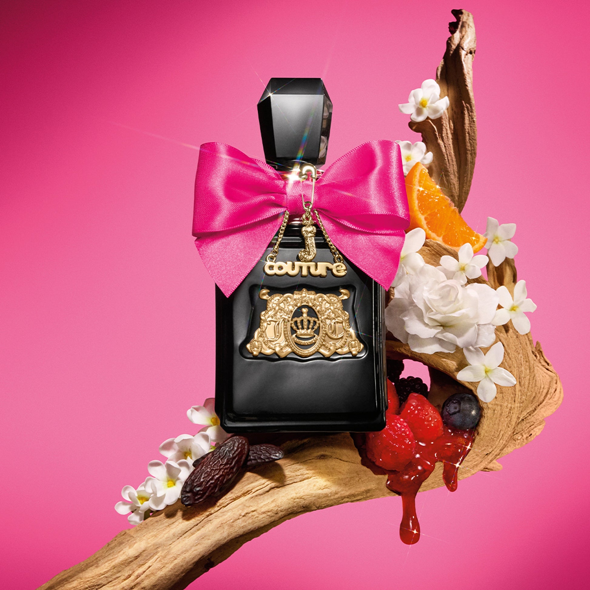 A Viva La Juicy Elixir Eau de Parfum Spray Pen, 0.33 oz, with black and gold accents and a pink bow rests on a wood branch amid white flowers, orange slices, and berries—a vibrant women’s fragrance by Juicy Couture.