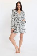 A woman poses barefoot in a Hooded Luxe Plush Robe with black cursive writing, front pockets, and a matching belt against a white background—an ideal pick for comfort and self-care.