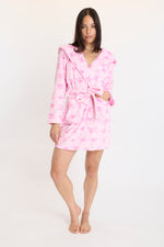 A woman with long dark hair stands barefoot in the Hooded Luxe Plush Robe, featuring a belted waist, pockets, and a subtle pattern. She faces the camera on a plain white background, embodying self-care and cozy comfort.