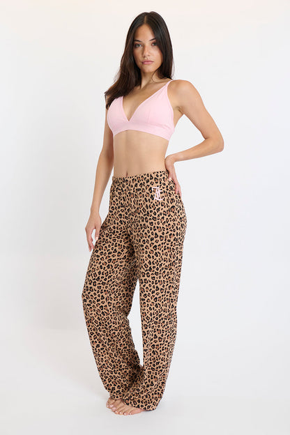 A woman with long dark hair and a neutral expression stands barefoot against a plain background, modeling the Plush 3-Piece PJ Set, which features a light pink triangle bralette and loose, high-waisted leopard print pants.
