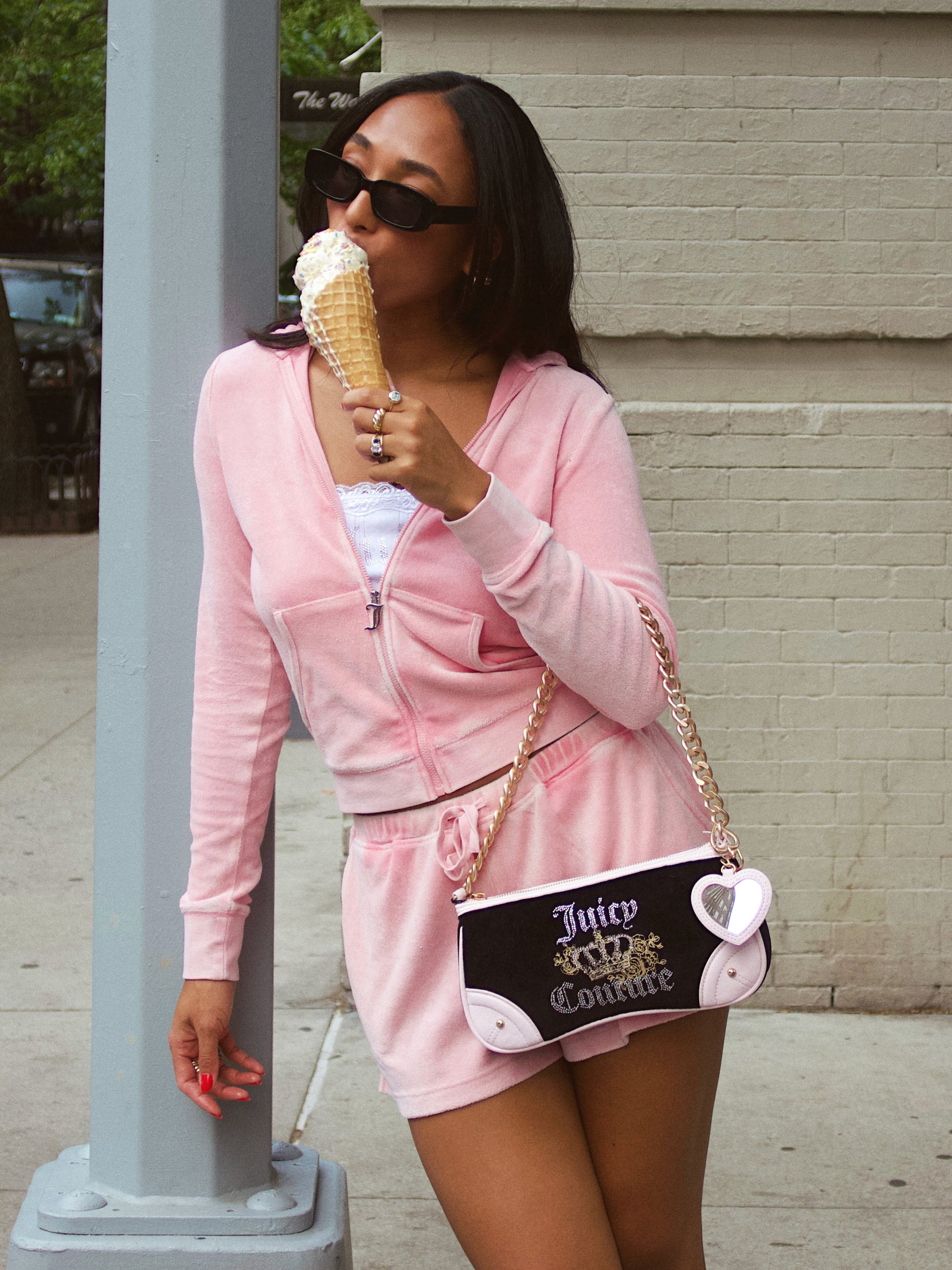 A woman in a pink velour tracksuit and sunglasses eats an ice cream cone on a city sidewalk, carrying the Royal Bloom Shoulder Bag with a heart charm that adds timeless glamour to her look.