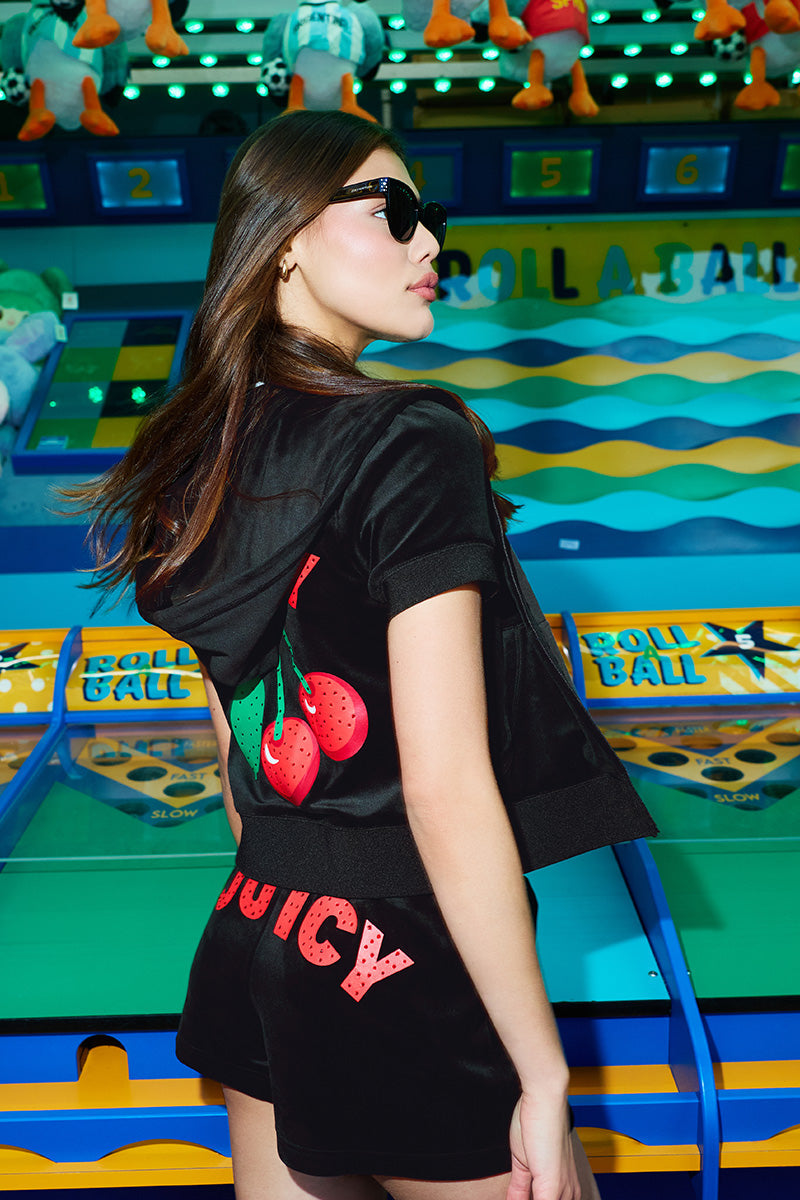 A woman in sunglasses and a black outfit with Cherry Bling Velour Shorts, featuring a cherry logo and JUICY graphic, stands before a colorful ROLL A BALL arcade game topped with plush toys.