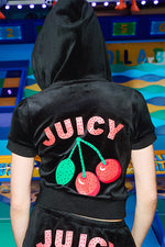 A person in the Cherry Bling Velour Hoodie, featuring a black velour short-sleeve design with a Juicy cherry graphic and "JUICY" on the back, stands before a vibrant arcade-like backdrop.