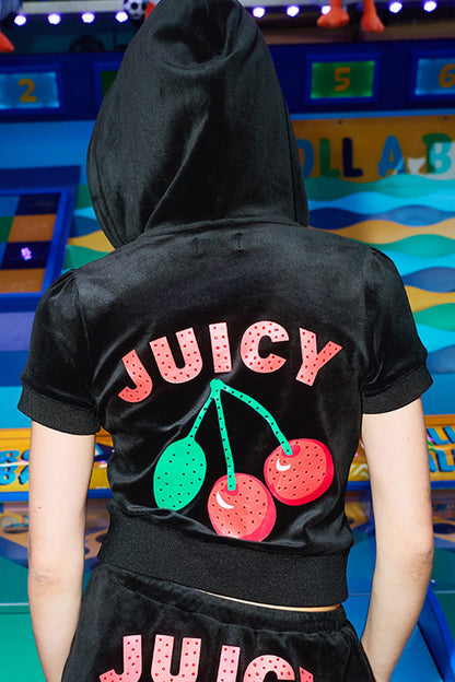 A person in the Cherry Bling Velour Hoodie, featuring a black velour short-sleeve design with a Juicy cherry graphic and "JUICY" on the back, stands before a vibrant arcade-like backdrop.