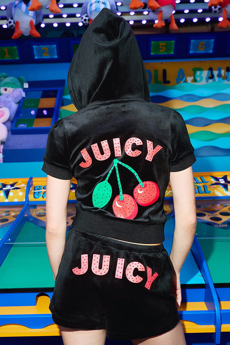 Someone wearing Cherry Bling Velour Shorts and a matching hoodie with a bold JUICY graphic and cherry logo on the back stands in front of a colorful carnival game booth.