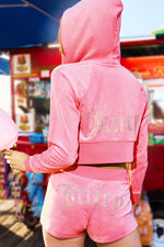 A person wearing the Scottie Big Bling Velour Hoodie in pink and matching shorts with rhinestone Juicy logos stands outdoors near vibrant vendor stalls.