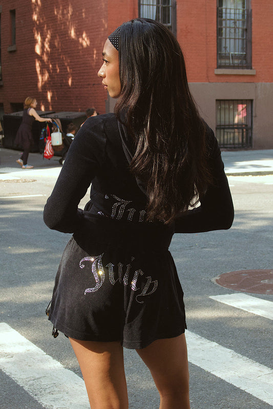 A woman with long dark hair, wearing a black headband and Big Bling Washed Terry Shorts featuring a rhinestone logo and drawstring waistband, stands at a city crosswalk facing away.