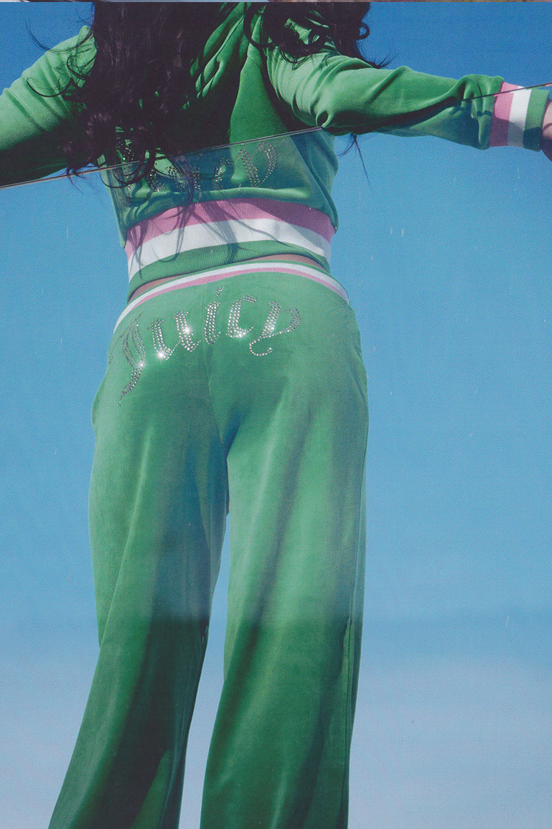 Wearing Heritage Stripe Bling Velour Track Pants with a rhinestone Juicy logo and sporty striped waistband, a person stands under a bright blue sky, arms partially outstretched and head out of frame.
