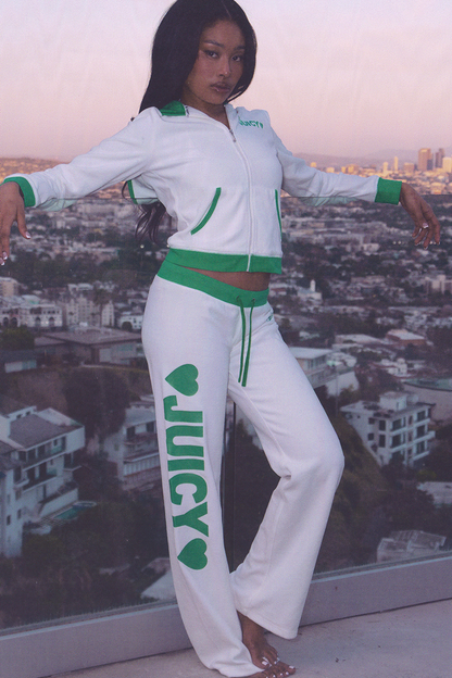 A woman stands on a balcony overlooking the city, wearing sporty loungewear—Contrast Low Rise Track Pants in white and green, featuring JUICY and heart motifs on her relaxed-fit pants.