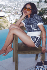 A woman in sunglasses and Logo Stripe Dolphin Booty Shorts sits barefoot on a poolside chair against a city backdrop, her face resting on her hand. A stylish handbag and relaxed vibe complete her chic look.