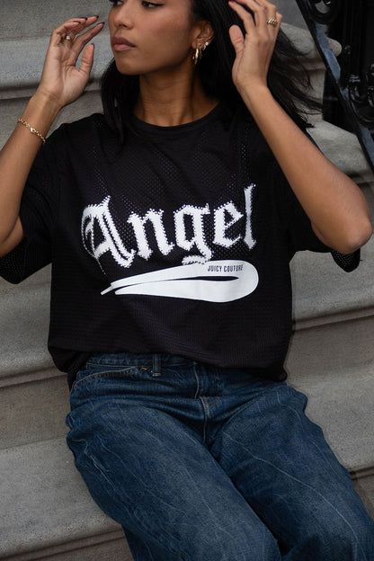 A woman sits on outdoor steps wearing the Angel Couture Oversized Tee Dress, featuring "Angel" in bold white gothic letters. She pairs it with blue jeans and adjusts her hair, radiating confidence.